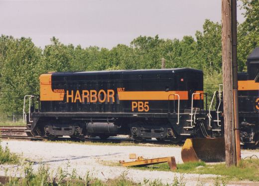Side view of a black cabless locomotive unit with an orange stripe and the lettering INDIANA HARBOR BELT and PB5, parked on a yard track with trees behind it and a rusty bucket and a yellow steel beam on the gravel in front.