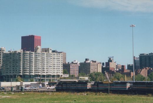 Three silver Amtrak diesel locomotives with red, white and blue stripes in a rail yard beyond a grassy field, with curved concrete apartment towers and downtown buildings under a blue sky.