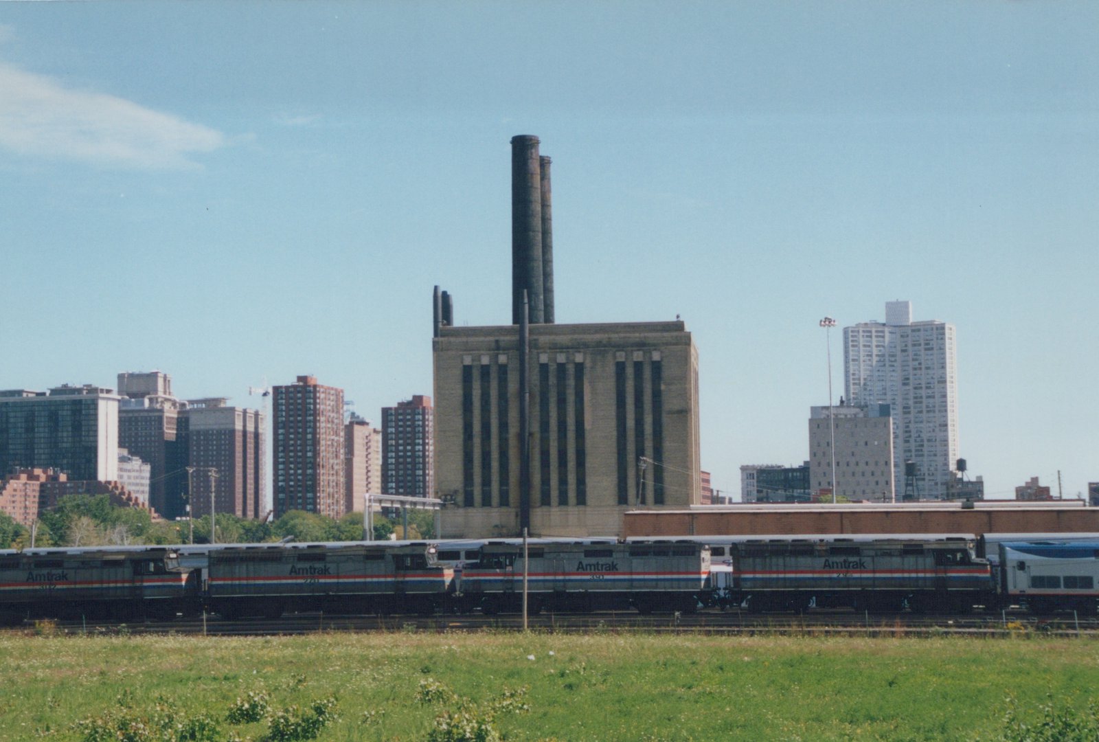 Four silver Amtrak diesel locomotives with red, white and blue stripes stand in a row in a rail yard beyond a grassy field, with a tall brick power house with two smokestacks and high-rise buildings behind them.