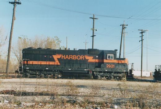 Side view of a black diesel locomotive with an orange stripe and large orange HARBOR lettering, number 2921 on the cab, standing on gravel-covered tracks under utility poles and a blue sky.