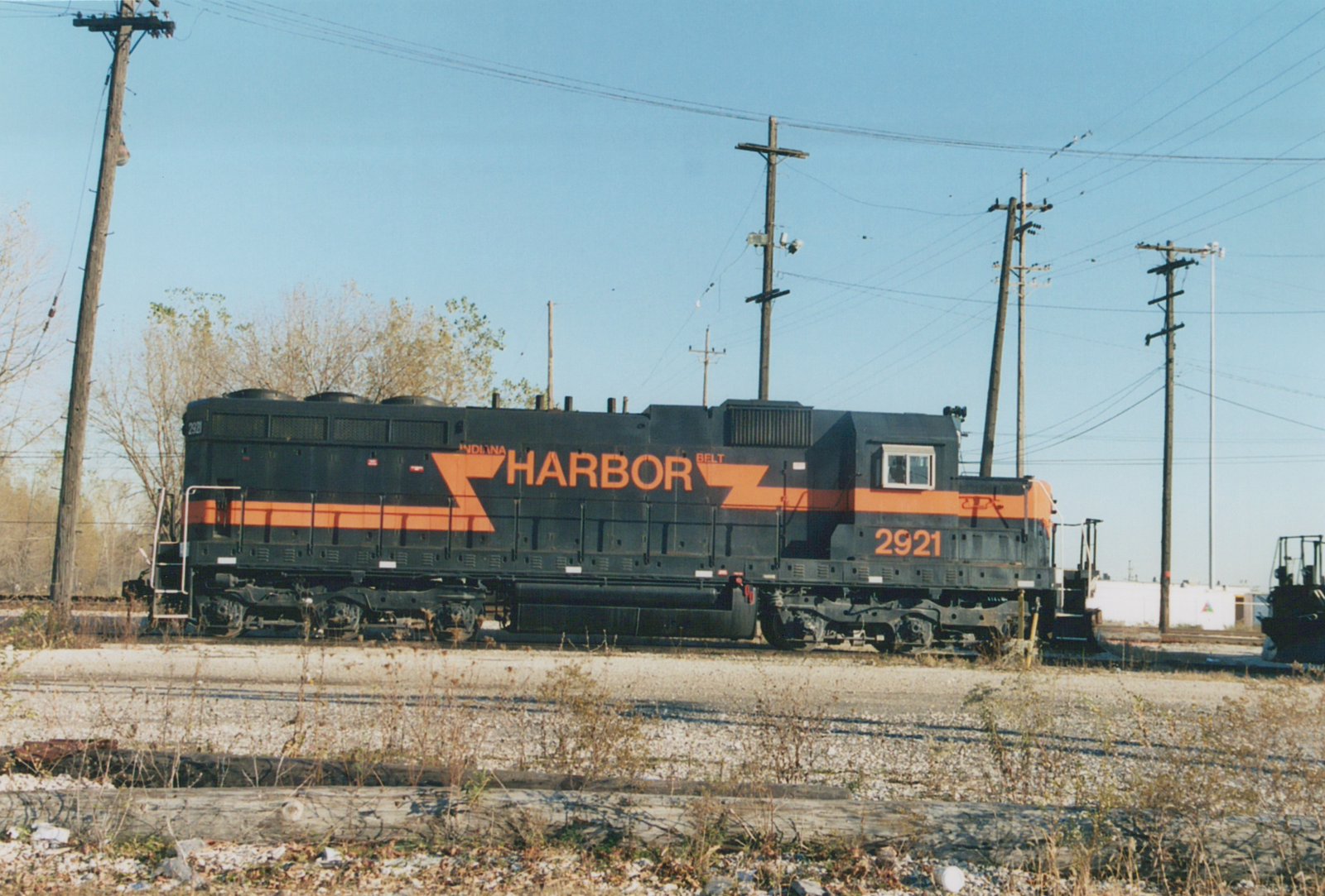 Side view of a black diesel locomotive with an orange stripe and large orange HARBOR lettering, number 2921 on the cab, standing on gravel-covered tracks under utility poles and a blue sky.