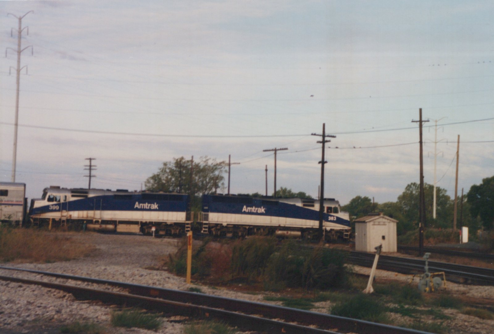 Two blue, white and silver Amtrak diesel locomotives numbered 399 and 383 coupled back to back with a silver passenger car at the left, seen across weedy tracks with a small white shed and utility poles.