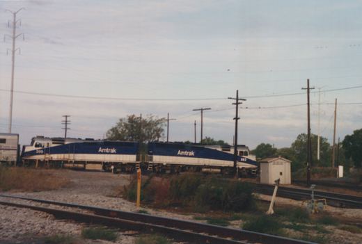 Two blue, white and silver Amtrak diesel locomotives numbered 399 and 383 coupled back to back with a silver passenger car at the left, seen across weedy tracks with a small white shed and utility poles.