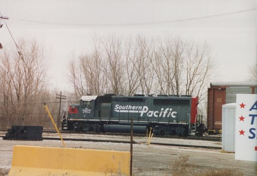 A gray locomotive with a red nose and large white Southern Pacific lettering stands on yard tracks, coupled to an autorack, with bare trees behind and a yellow concrete barrier in the foreground.