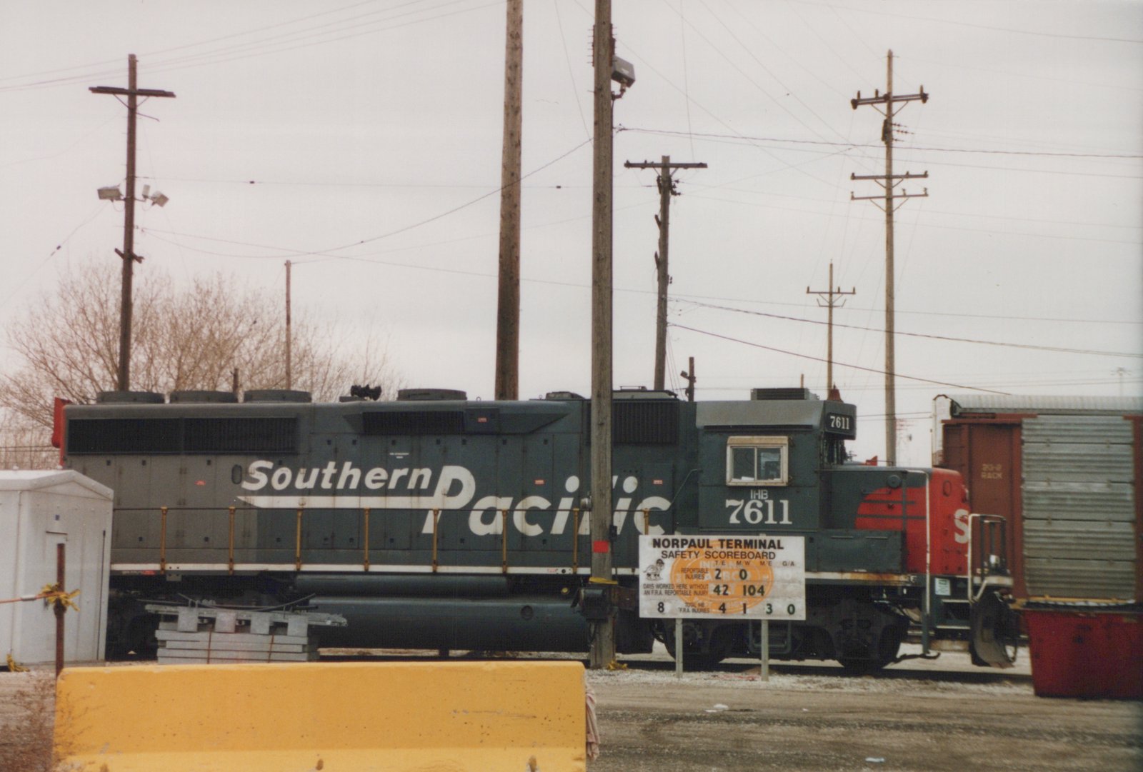 A dark gray diesel locomotive with a red nose and large white Southern Pacific lettering stands in a rail yard behind a safety scoreboard sign and a yellow concrete barrier, under an overcast sky.