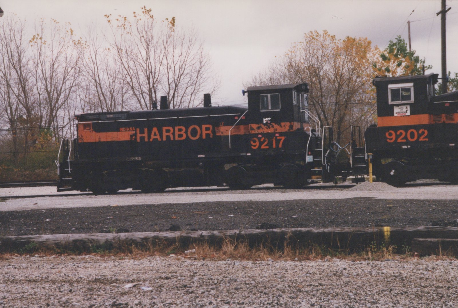A black diesel switcher with an orange stripe and HARBOR in large orange letters, numbered 9217, is coupled to a second black and orange locomotive, numbered 9202, on gravel yard tracks with autumn trees behind.