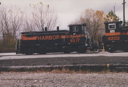 A black diesel switcher with an orange stripe and HARBOR in large orange letters, numbered 9217, is coupled to a second black and orange locomotive, numbered 9202, on gravel yard tracks with autumn trees behind.