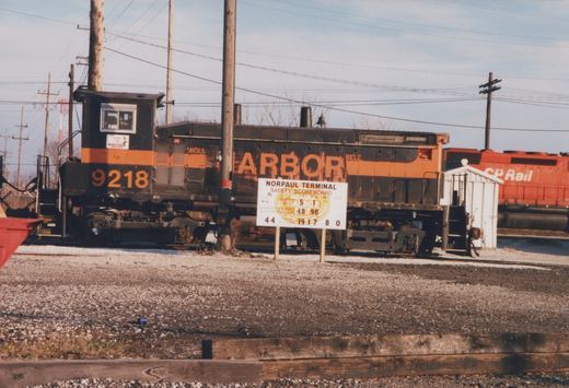 A weathered black and orange switcher locomotive numbered 9218 stands on a gravel-lined track behind a white safety sign, with a red CP Rail locomotive at the right.