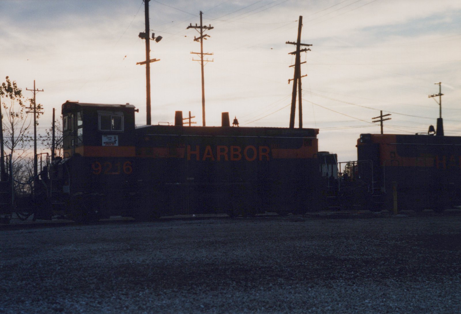 A black and orange switcher locomotive lettered HARBOR stands against a dusk sky beneath utility poles, with a second locomotive behind it at right and a gravel lot in the foreground.