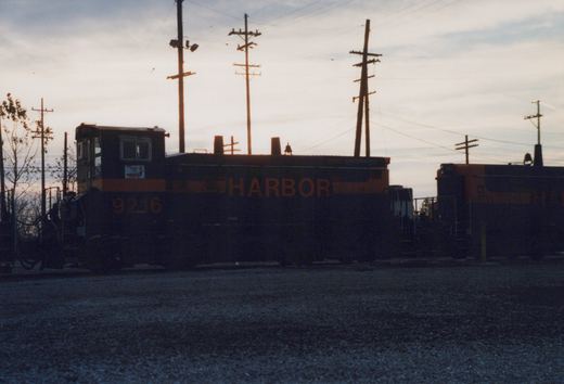 A black and orange switcher locomotive lettered HARBOR stands against a dusk sky beneath utility poles, with a second locomotive behind it at right and a gravel lot in the foreground.