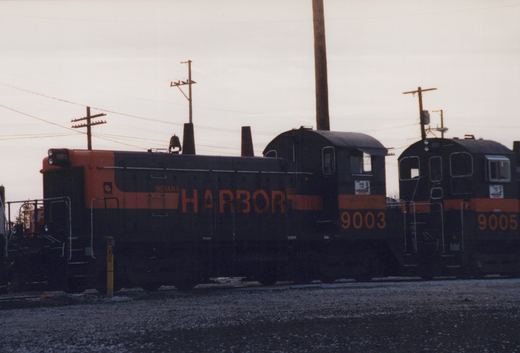 Side view of a black end-cab switcher with an orange hood end, lettered INDIANA HARBOR BELT and numbered 9003, coupled cab to cab with another black and orange switcher under an overcast sky.