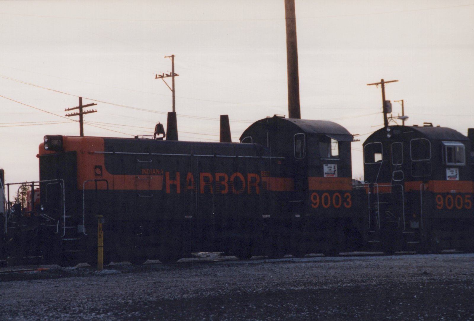 Side view of a black end-cab switcher with an orange hood end, lettered INDIANA HARBOR BELT and numbered 9003, coupled cab to cab with another black and orange switcher under an overcast sky.