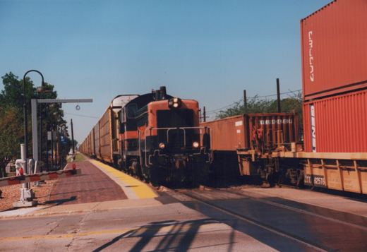 Head-on view of an orange-and-black switcher locomotive with its headlights on, pulling a long line of yellow autorack cars beside a brick station platform, with lowered crossing gates at left and red stacked containers on a well car at right.