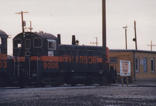 A black diesel switcher with an orange stripe, lettered Harbor and numbered 9005, stands beside another switcher in a gravel yard in front of a yellow brick building, under an overcast sky.