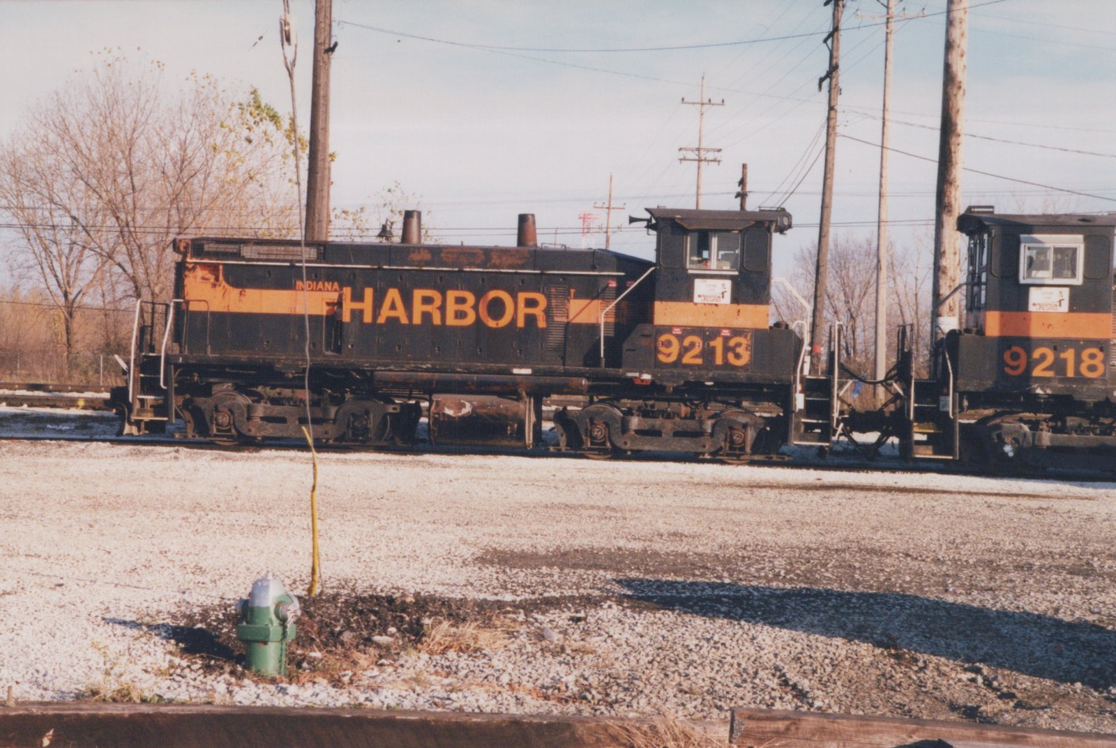 Side view of a weathered black and orange diesel switcher lettered INDIANA HARBOR and numbered 9213, coupled to a second unit numbered 9218, behind a gravel lot with a green fire hydrant.