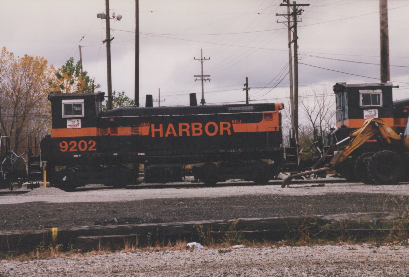 A black diesel switcher with an orange stripe, lettered HARBOR and numbered 9202, stands on a yard track, with a second switcher and a yellow front-end loader at right and utility poles against a grey sky.