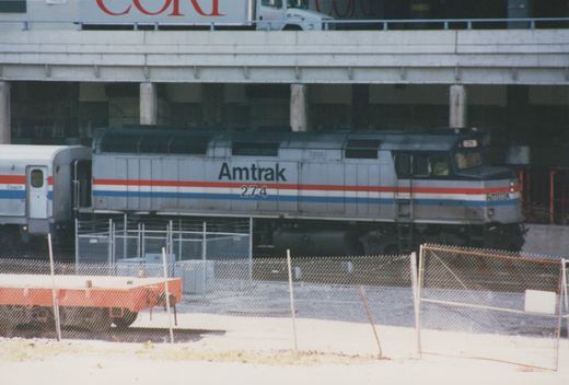 A silver Amtrak diesel locomotive numbered 274, with red, white and blue stripes, coupled to a coach beneath a concrete street viaduct, seen through a chain-link fence.