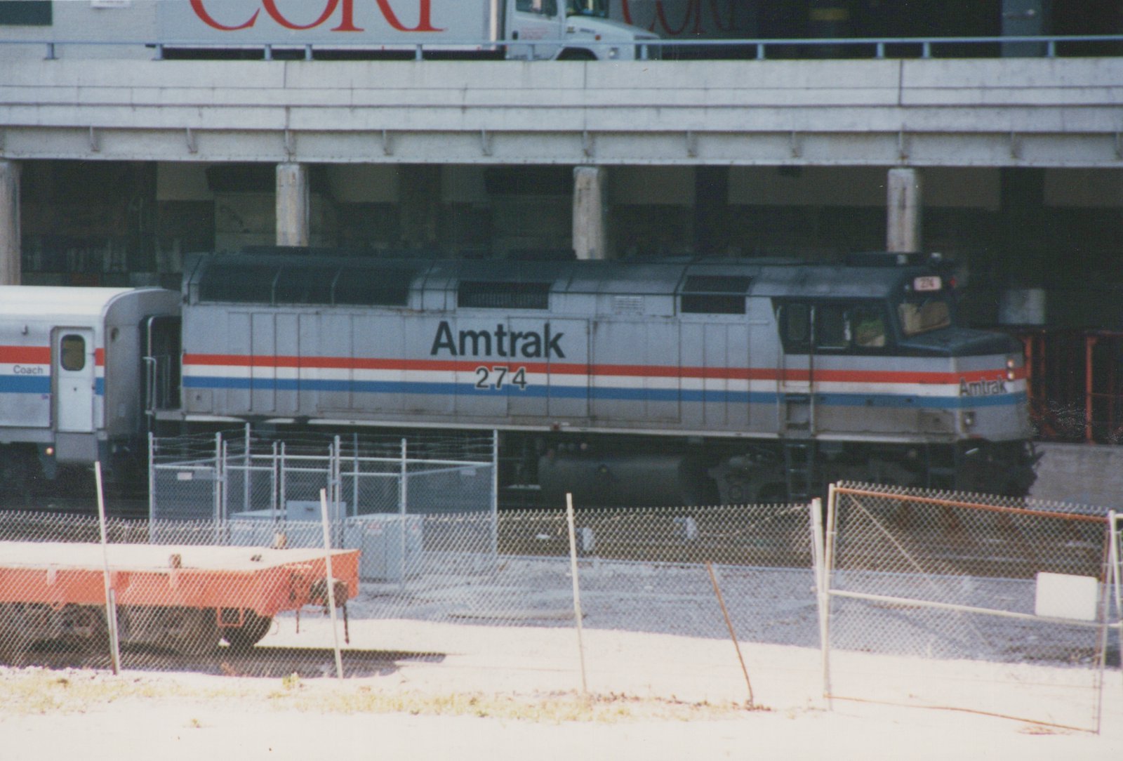 A silver Amtrak diesel locomotive numbered 274, with red, white and blue stripes, coupled to a coach beneath a concrete street viaduct, seen through a chain-link fence.
