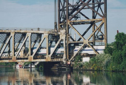 Two silver and blue Amtrak diesel locomotives seen through the rusted lattice truss of a river bridge with a tall lift tower, with moored boats and the still water in the foreground.