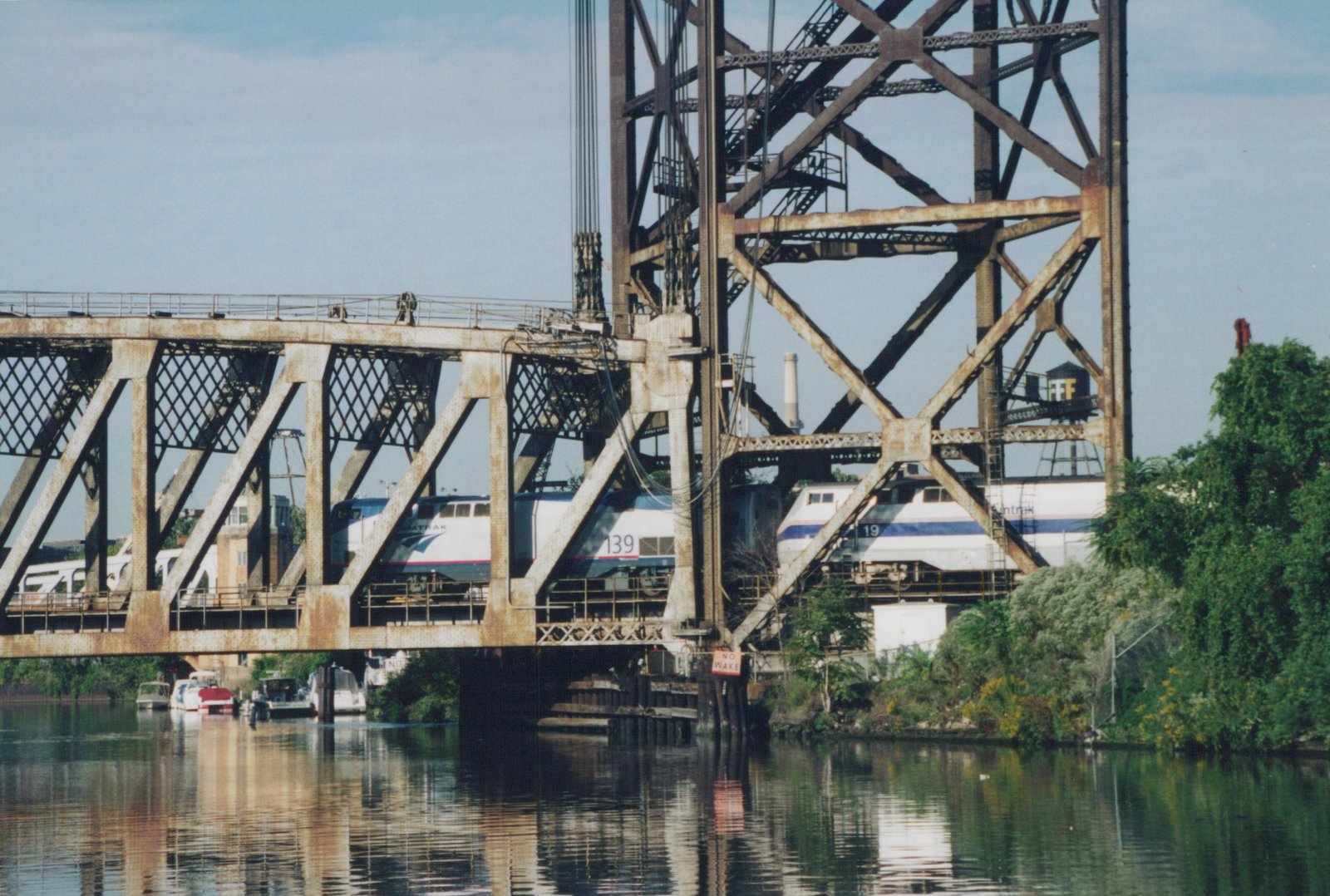Two silver and blue Amtrak diesel locomotives seen through the rusted lattice truss of a river bridge with a tall lift tower, with moored boats and the still water in the foreground.