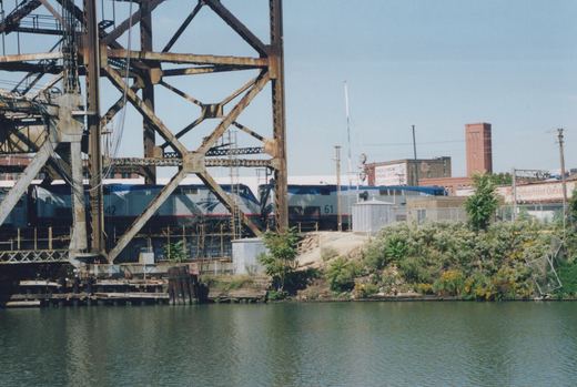 Two silver and blue Amtrak diesel locomotives, numbered 42 and 61, partly hidden behind a rusted steel bridge tower on the far bank of a green river lined with weeds and old brick buildings.