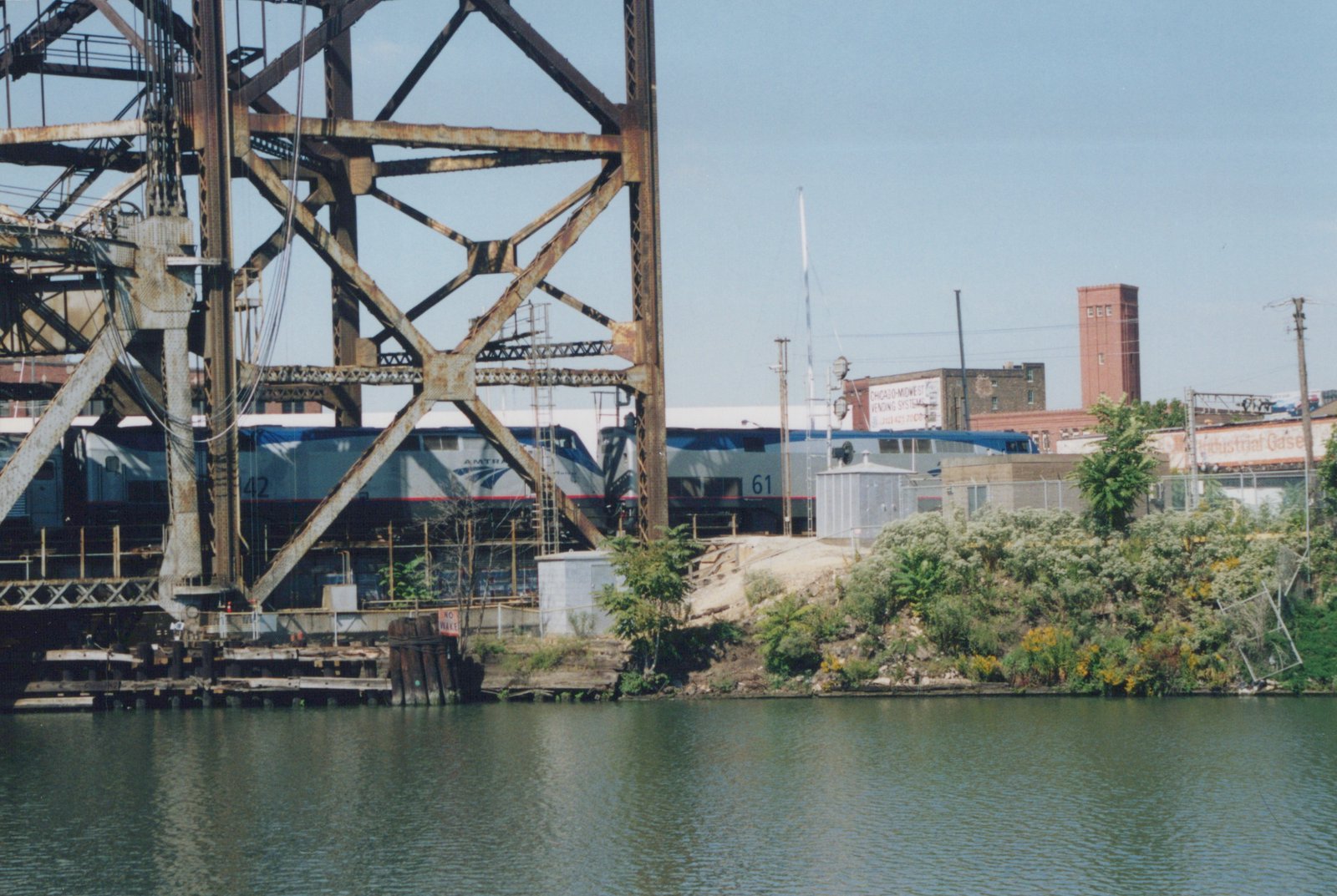 Two silver and blue Amtrak diesel locomotives, numbered 42 and 61, partly hidden behind a rusted steel bridge tower on the far bank of a green river lined with weeds and old brick buildings.