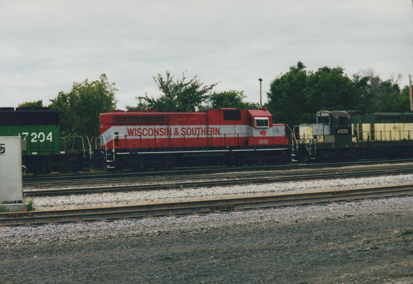 Side view of a red and white diesel locomotive lettered Wisconsin & Southern, number 2052, parked in a yard between a green locomotive numbered 7204 and a faded green and cream locomotive numbered 6622, under an overcast sky.