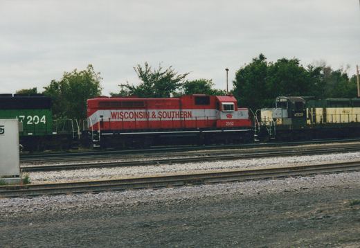 Side view of a red and white diesel locomotive lettered Wisconsin & Southern, number 2052, parked in a yard between a green locomotive numbered 7204 and a faded green and cream locomotive numbered 6622, under an overcast sky.