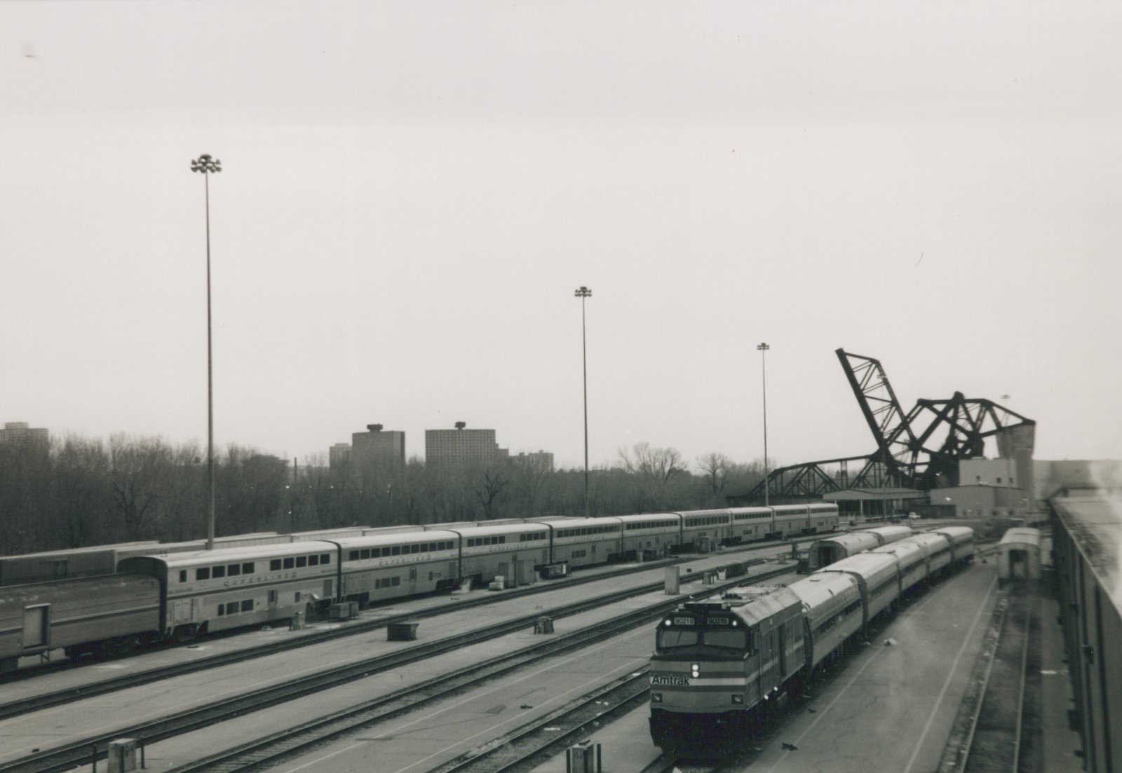 Black-and-white photo looking down on a railroad yard. An Amtrak cab unit numbered 90219 leads a short train of single-level cars. Long bilevel Superliner cars stand on tracks to the left, with tall light towers, leafless trees, high-rise buildings and a raised bascule bridge behind.