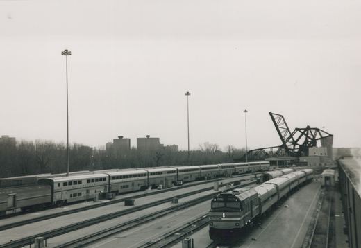 Black-and-white photo looking down on a railroad yard. An Amtrak cab unit numbered 90219 leads a short train of single-level cars. Long bilevel Superliner cars stand on tracks to the left, with tall light towers, leafless trees, high-rise buildings and a raised bascule bridge behind.