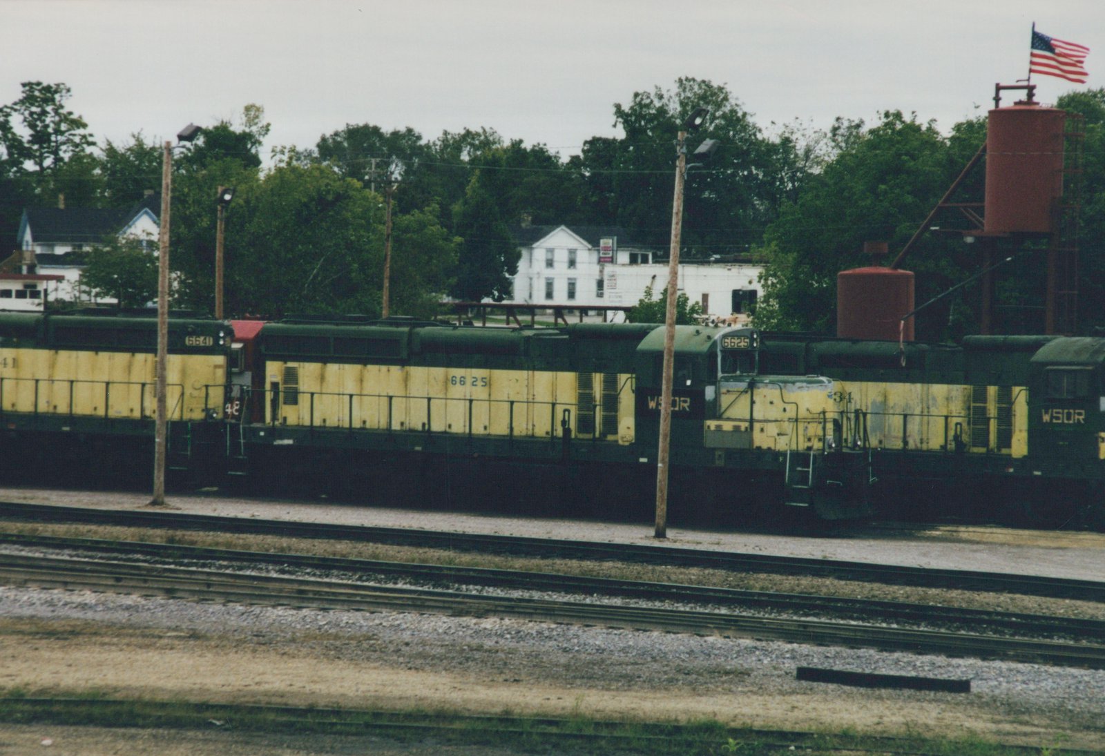 A line of weathered yellow and dark green diesel locomotives, two lettered WSOR, parked beside yard tracks under an overcast sky, with red water tanks, an American flag and white houses behind.