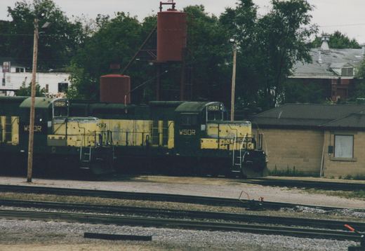 Three diesel locomotives in faded yellow and dark green with WSOR on the cab sides stand in a row beside two red tanks on a steel frame, with yard tracks in the foreground and trees and buildings behind.