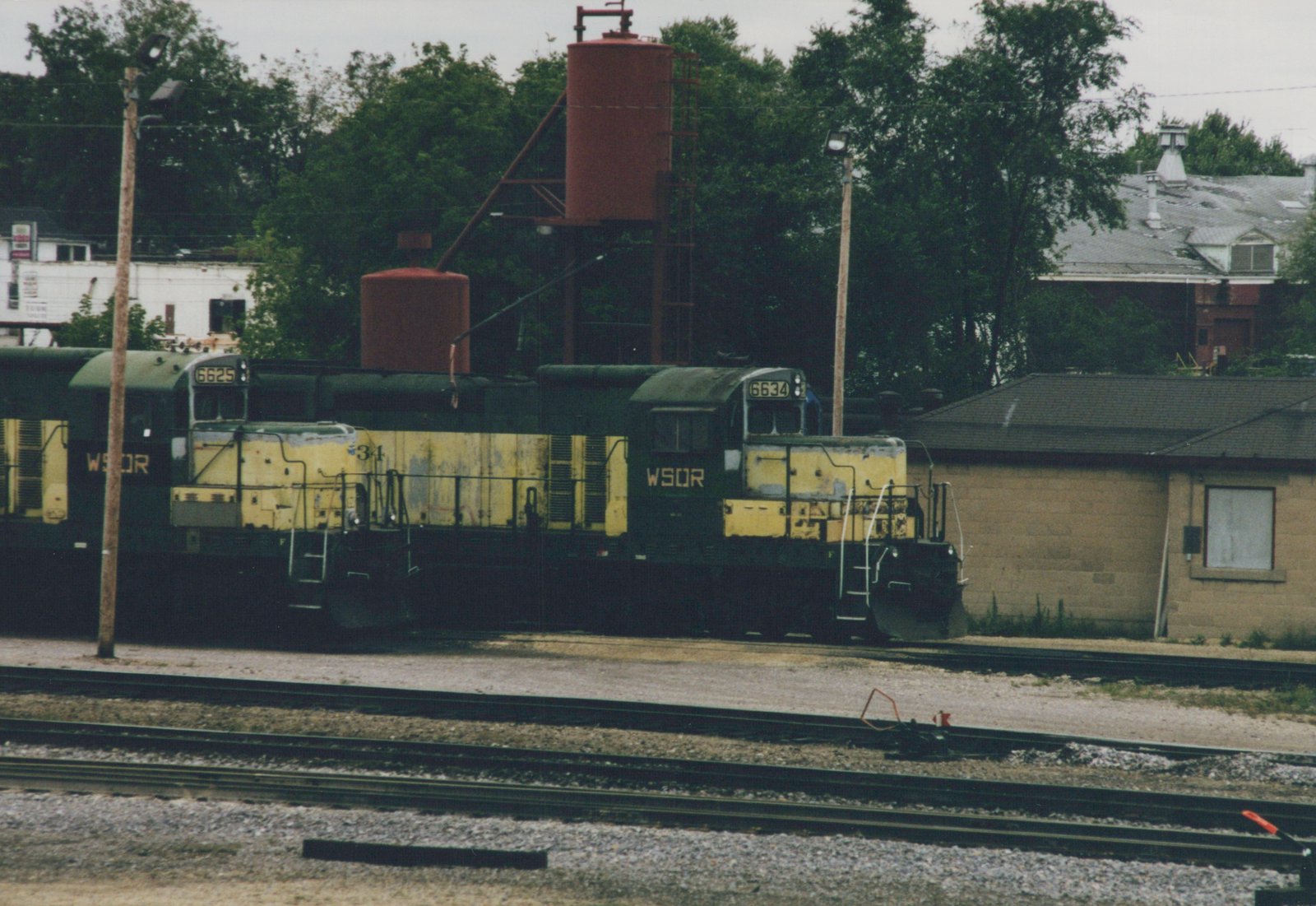 Three diesel locomotives in faded yellow and dark green with WSOR on the cab sides stand in a row beside two red tanks on a steel frame, with yard tracks in the foreground and trees and buildings behind.