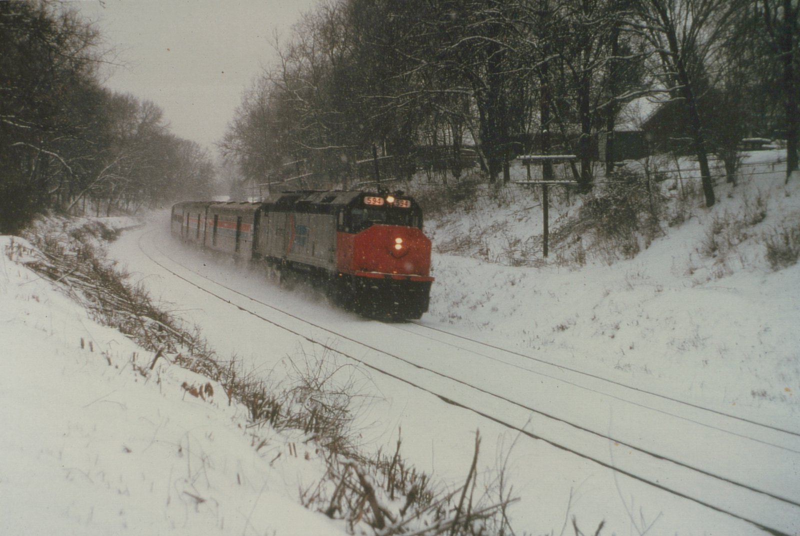 A red-nosed, silver Amtrak diesel locomotive with its headlights on pulls a short train of stainless steel passenger cars around a snow-covered curve between wooded banks.
