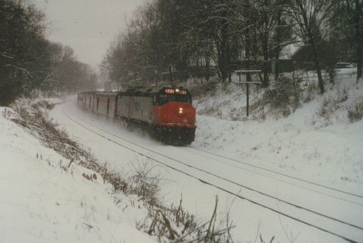 A red-nosed, silver Amtrak diesel locomotive with its headlights on pulls a short train of stainless steel passenger cars around a snow-covered curve between wooded banks.