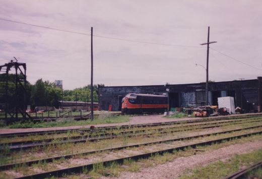 An orange-nosed brown diesel locomotive stands outside a dark, weathered roundhouse with scrap piled nearby, seen across grass-grown tracks under an overcast sky.
