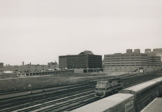 Black-and-white photo taken from above of a striped diesel locomotive numbered 516 on a broad spread of tracks, with bilevel passenger cars in the foreground and office buildings beyond a vacant lot.