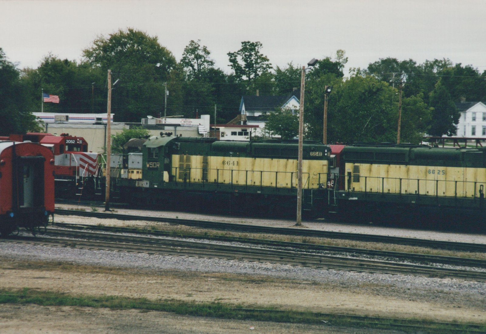 Two green and yellow diesel locomotives numbered 6641 and 6625 on a yard track, with a red and white locomotive numbered 2052 and the red end of a passenger car at left, trees and houses behind.