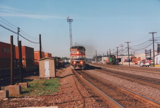 A silver locomotive with red, white and blue bands and its headlights lit approaches head-on along a ballasted track, with stacked orange containers on the left, a green switcher and industrial buildings on the right, and a tall light tower above.