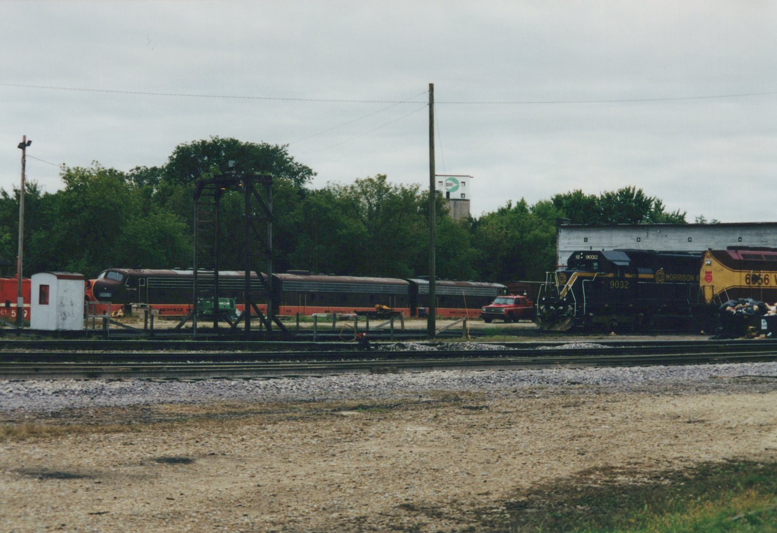 A black locomotive with yellow stripes numbered 9032 stands beside a white engine house, next to a yellow and maroon locomotive, while a maroon cab unit and two passenger cars with orange bands sit behind a fence under an overcast sky.