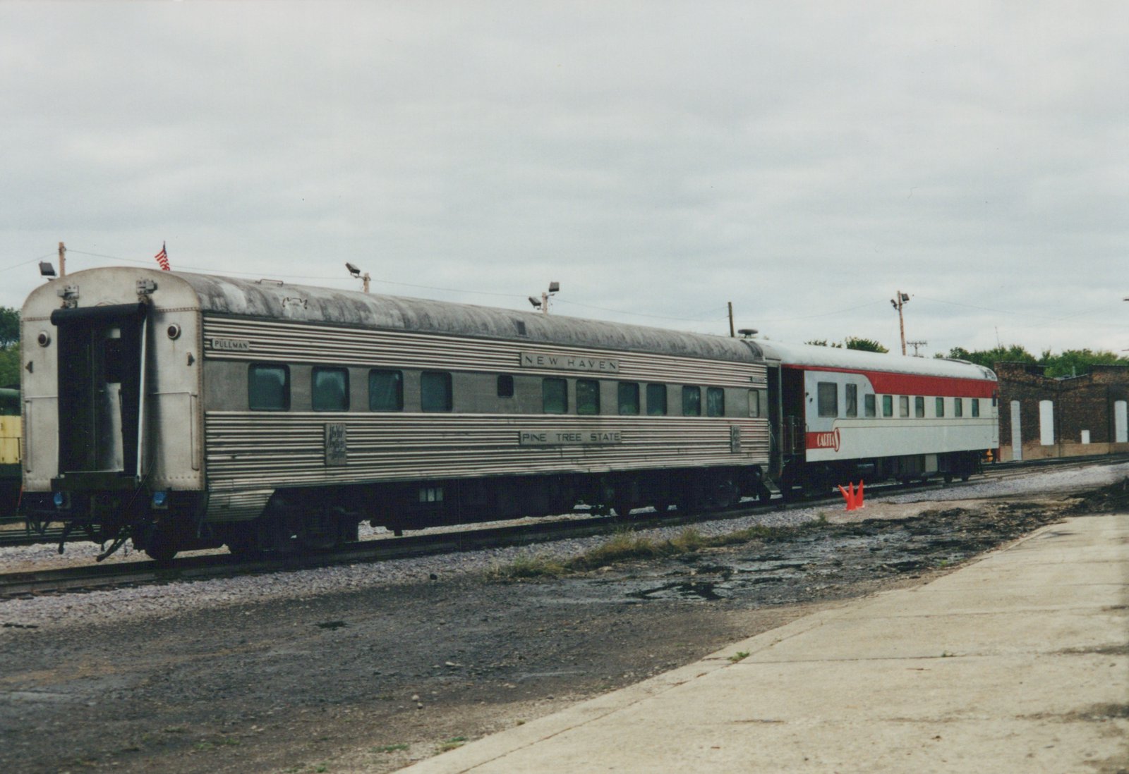 A fluted stainless steel passenger car lettered NEW HAVEN and PINE TREE STATE, seen from the rear corner, coupled to a white car with a red stripe, in front of a brick building under a cloudy sky.