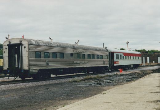 A fluted stainless steel passenger car lettered NEW HAVEN and PINE TREE STATE, seen from the rear corner, coupled to a white car with a red stripe, in front of a brick building under a cloudy sky.