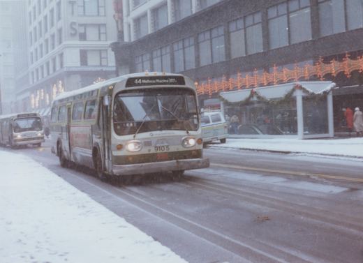 A white and green Chicago Transit Authority bus drives down a snow-covered city street with holiday lights in the background.