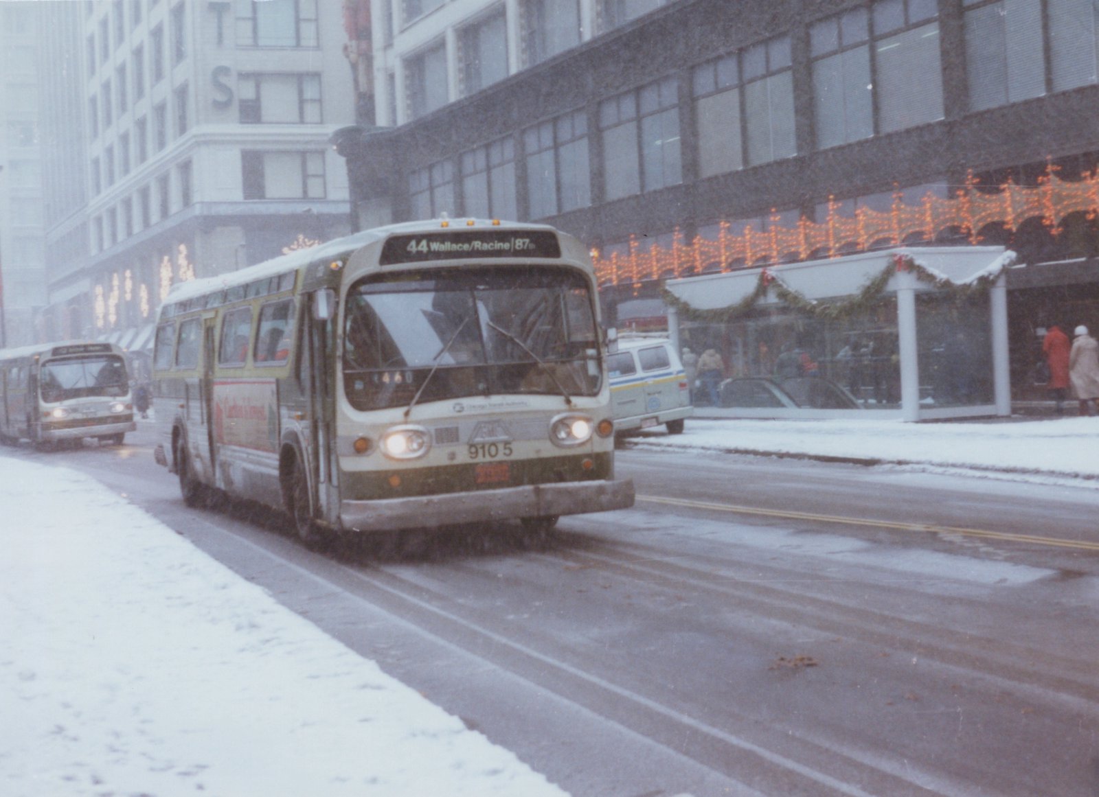 A white and green Chicago Transit Authority bus drives down a snow-covered city street with holiday lights in the background.