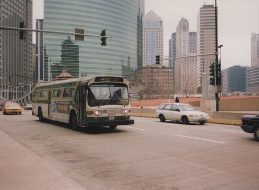 A green and cream Chicago Transit Authority motor bus numbered 9054 is driving on a wide city street, with tall glass and steel skyscrapers behind it and other cars nearby.