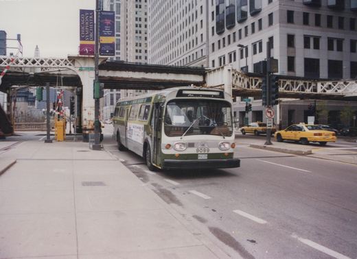 A green and cream CTA bus, number 9099, on a city street under an elevated train track, with yellow taxis in the background.