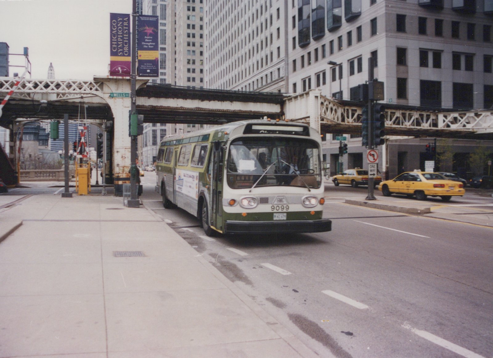 A green and cream CTA bus, number 9099, on a city street under an elevated train track, with yellow taxis in the background.