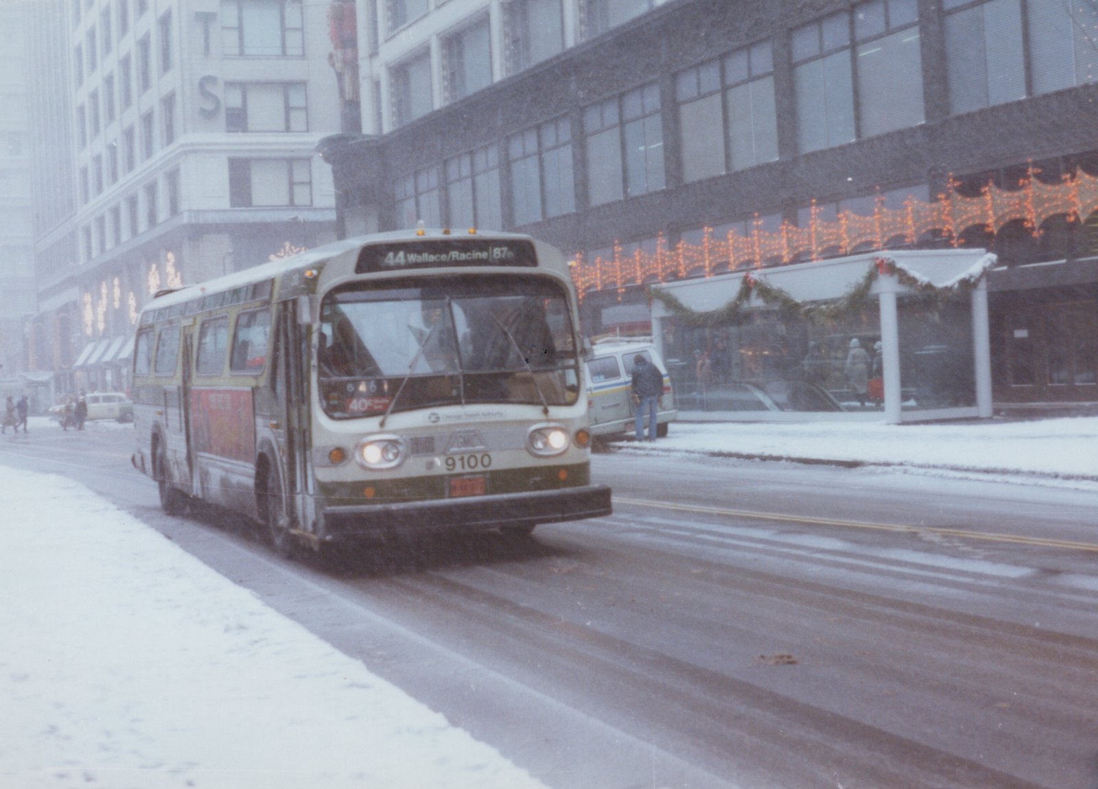 A cream-colored CTA bus, number 9100, drives on a snow-covered city street, passing buildings decorated with Christmas lights.