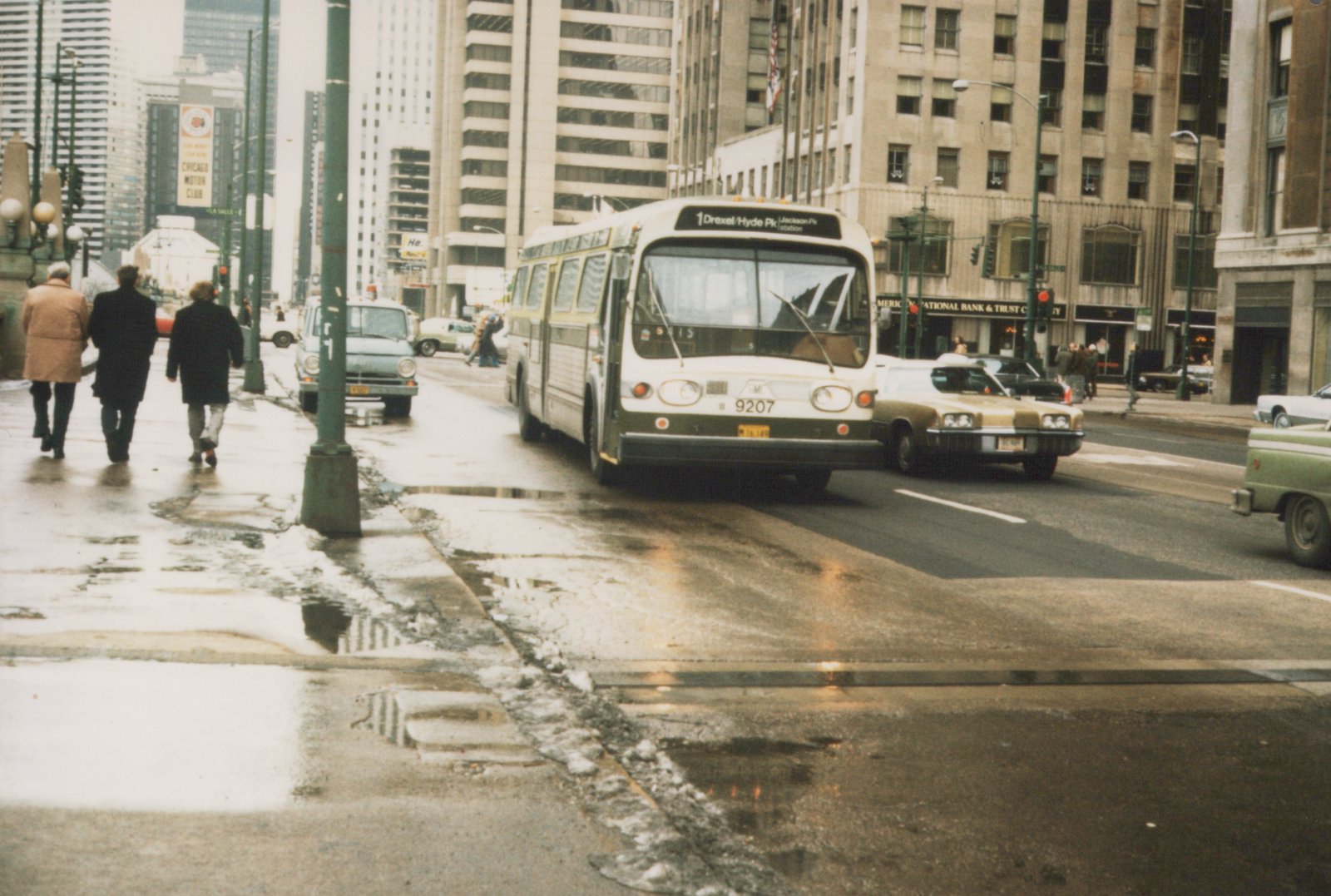 A white and green CTA bus labeled '9207' is stopped on a wet city street, surrounded by 1970s cars and pedestrians in winter coats, with tall buildings in the background.
