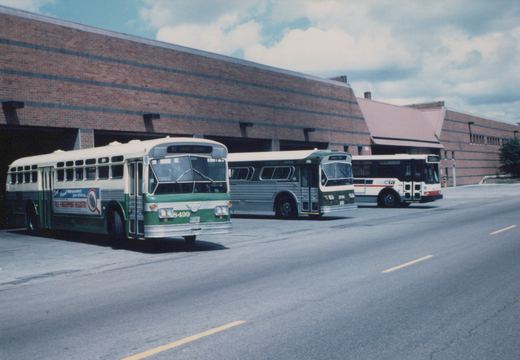 Three CTA buses, one green and two in white or dark green livery, parked in front of a brick building with garage doors.